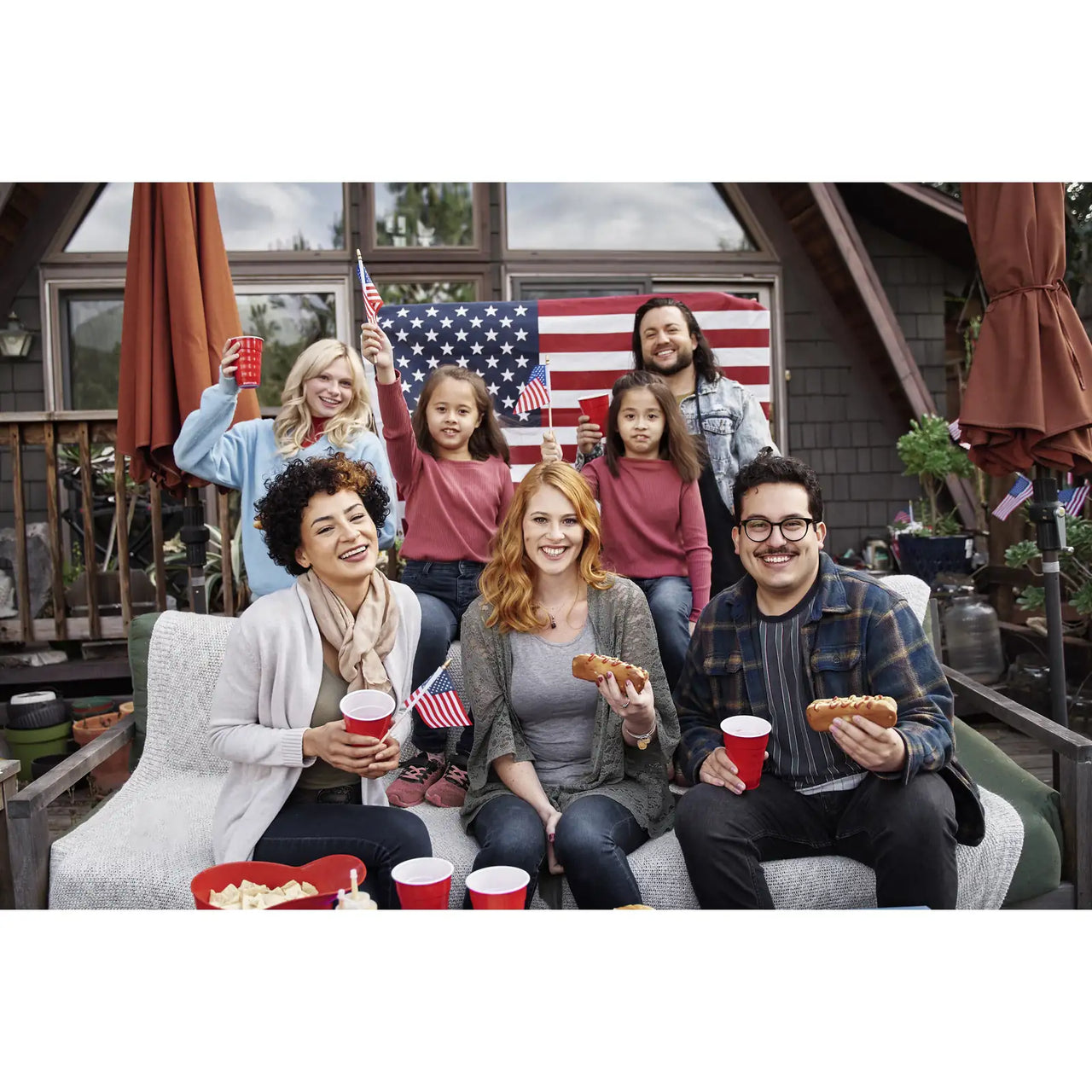 4th of July Photo Puzzle landscape orientation of a group of seven people sitting and standing together on a patio in front of a house, holding small American flags and red cups, with a large American flag displayed behind them celebrating 4th of july