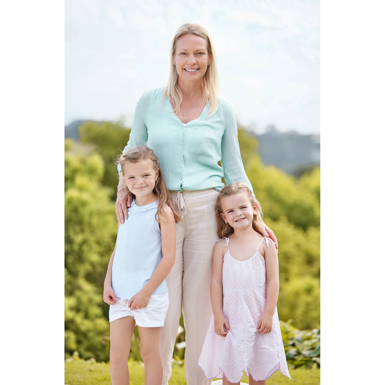 Personalized Mom Puzzle of a young mom standing outdoors with her two young girls in light blue and pale pink sleeveless dresses, wearing a mint green button-front cardigan and beige pants.